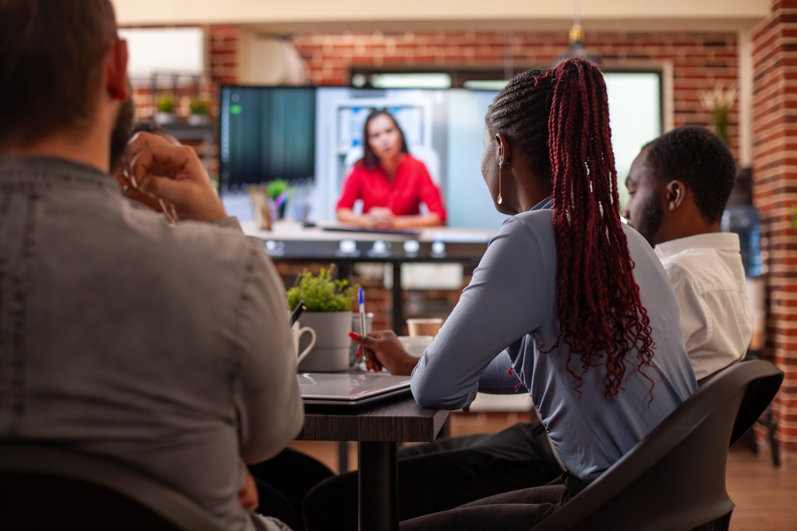 Black woman looks at video call on screen Black woman and coworkers sit at table, attentively listening to client on video call displayed on monitor. Selective focus on african american lady looking at screen showing virtual call with employer